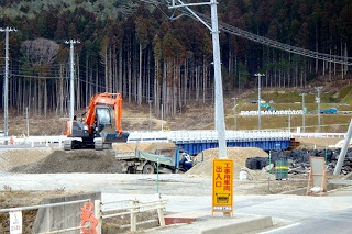 ショベルカーが土を掘り起こしてダンプカーに積み込んでおり、後方には森と山が見える写真