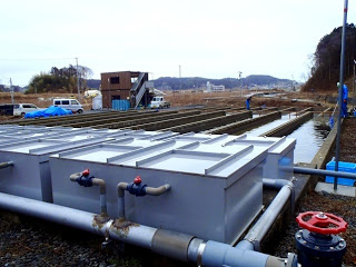 地面は砂利と土に覆われ、手前には金属製の大きなタンクや水槽が複数並びそれぞれ蓋が付いた四角い箱型の形をしている屋外に設置された養殖場の施設設備の写真