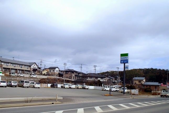 曇り空の下、住宅街の前にあるコンビニ「ファミリーマート」と広い駐車場が写された写真