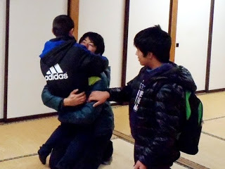 畳の部屋にいる三人の人物がいて、一人は膝をついて座り、アディダスのジャケットを着た子どもを抱きしめもう一人はその近くに立っており、膝をついた人物の肩に手を添えている写真