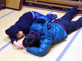 畳の上に2名の人が並んで寝そべっており、顔を伏せている人の手を左の人が触ろうとしている写真