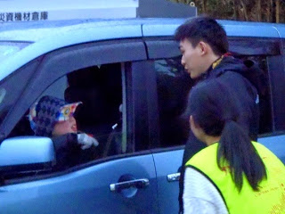 車の中の人に男性が声掛けしている様子を隣で女性が見ている写真