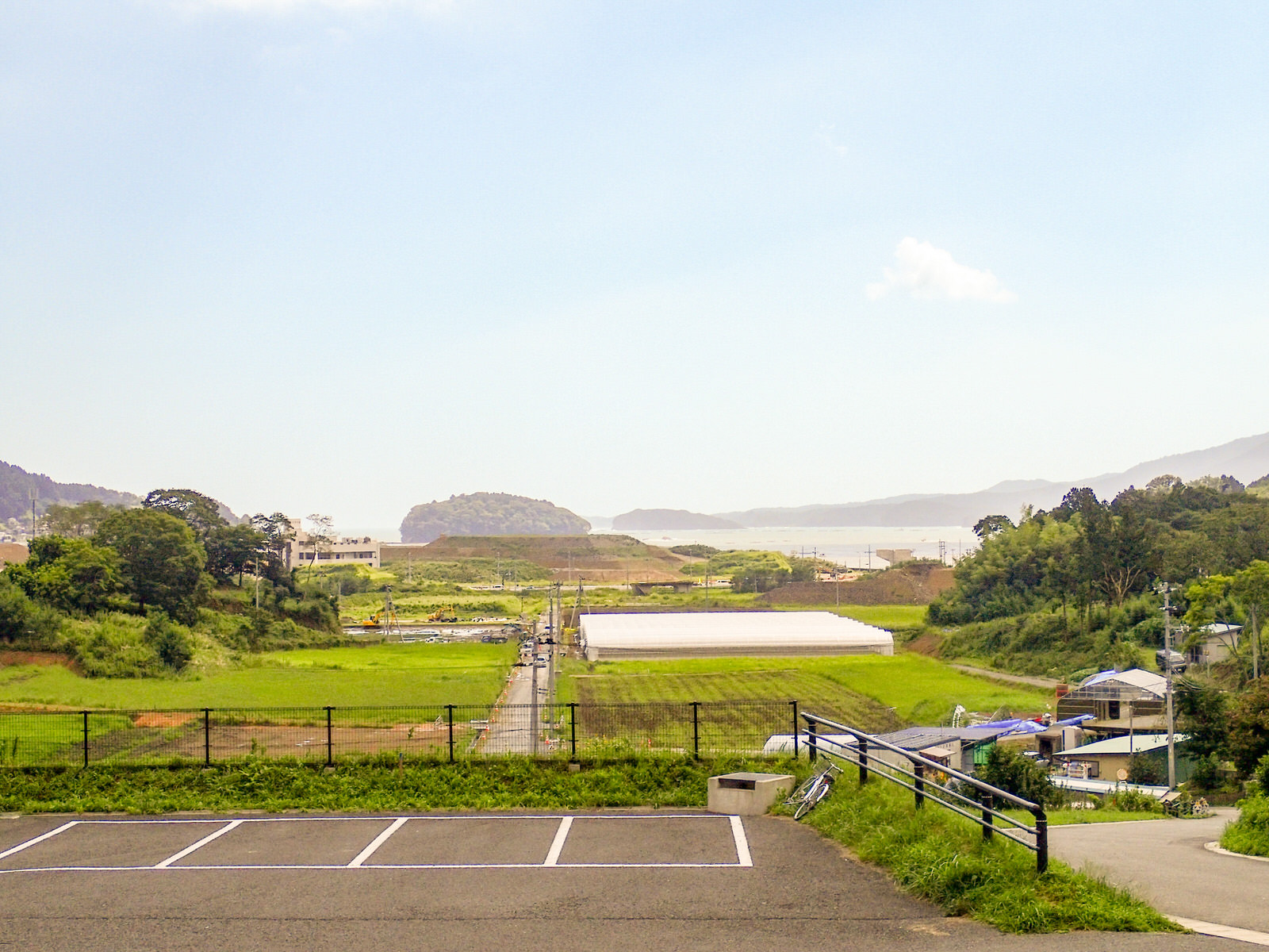 小高い山々と遠くに海が見える手前に、ビニールハウスや水田が広がる自然に囲まれたのどかな田園風景を、アスファルトの駐車場がある園庭から撮影した写真