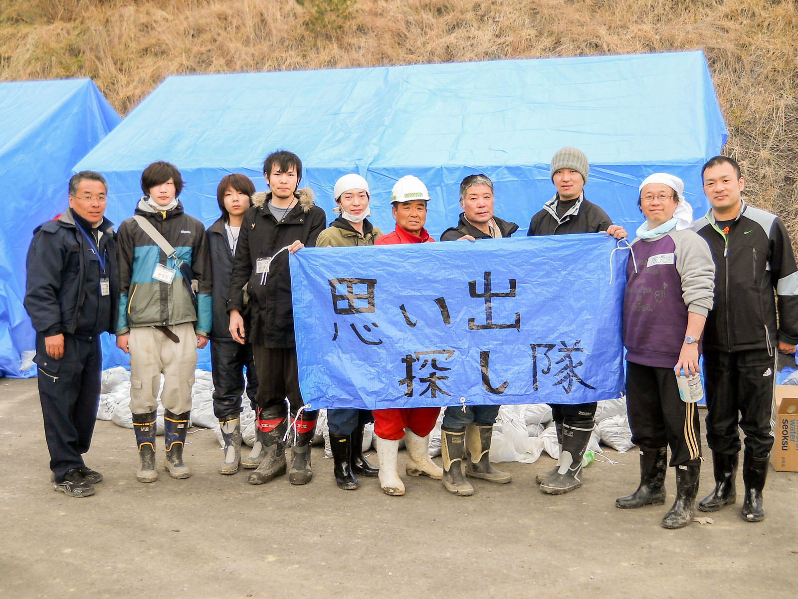 ブルーシートで覆われたテントの前で、「思い出探し隊」と手書きで書かれたブルーシートを持った10人のボランティアの方々が並んで立っている写真