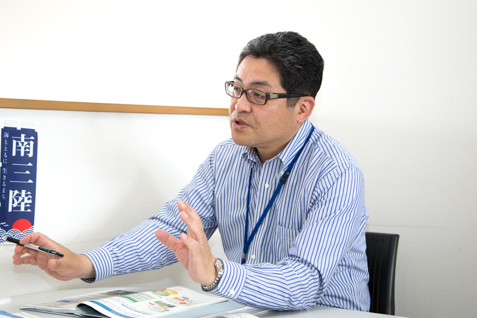 資料を広げた机に座った青と白のストライプ柄のシャツを着た千葉課長が話をしている様子の写真