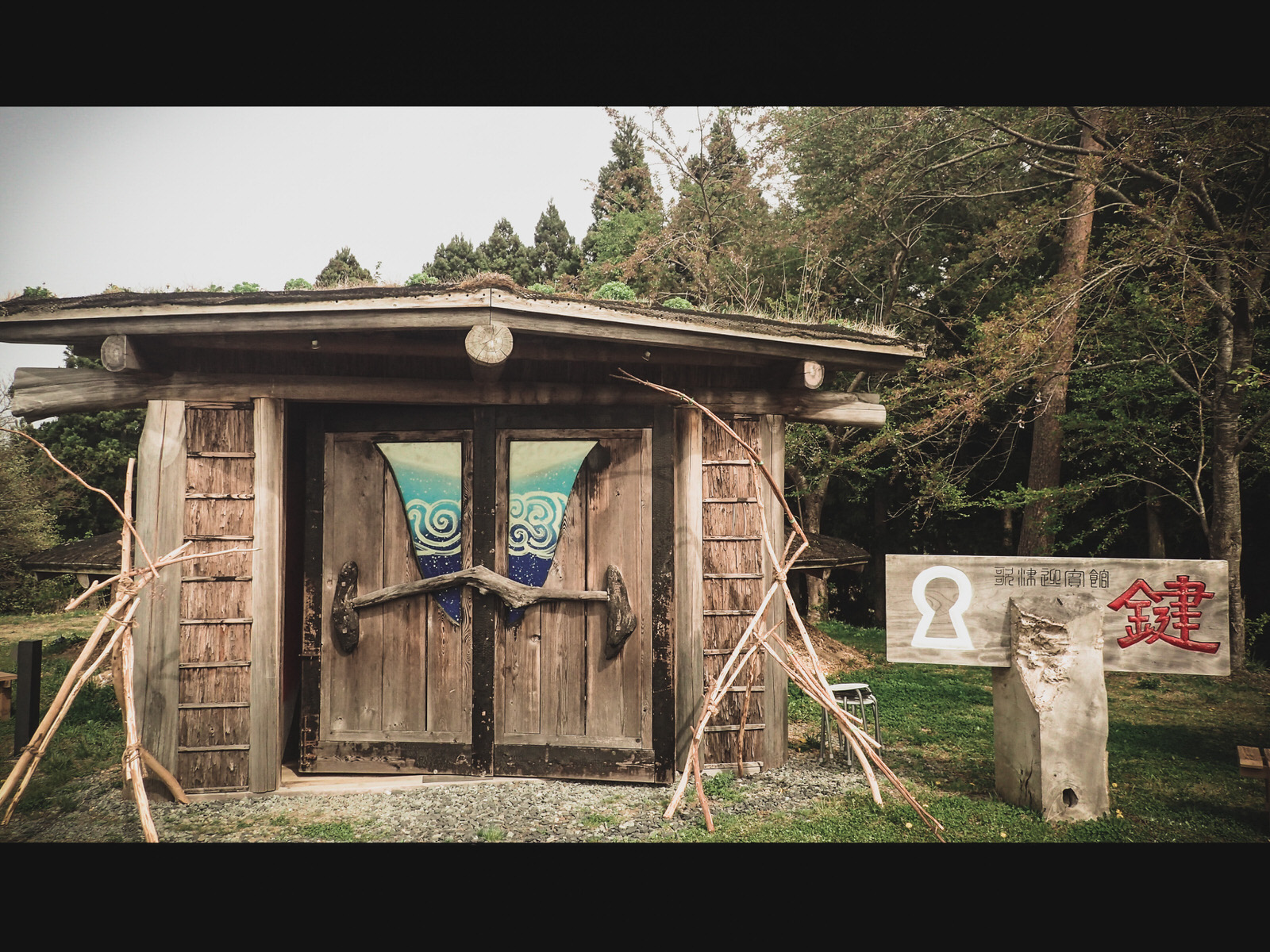 素朴な木造の小屋が林の中に建ち、扉には波模様がデザインされ、特徴的な木のドアノブがついている写真