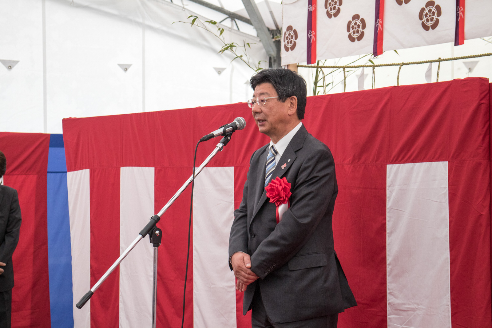 赤白幕の前でスーツ姿の佐藤町長がマイクスタンドに向かって真剣な表情でスピーチをしている写真