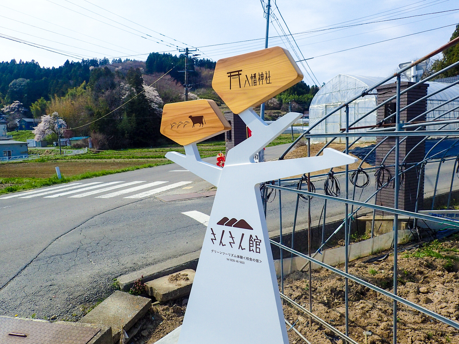 中央に「さんさん館」と書かれた、枝分かれした木のような形の白い土台の左側に、稲と牛のイラストが描かれた木の板、てっぺんに「八幡神社」と書かれた木の板がついており、右側が矢印の形になっている案内板が五叉路に設置されている写真