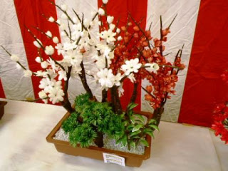 赤白の幕を背景に、机の上に置かれた長方形の鉢に白と赤の花が咲く枝ものと緑の葉が植えられているシルクフラワーの写真