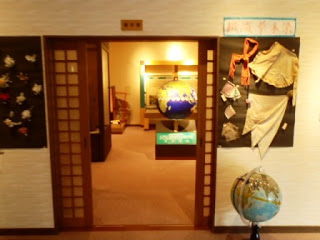 和風の木枠の引き戸が開かれた入口の奥に地球儀や展示物が見え、入口横の壁にはシルク工芸品が飾られた、展示ルームの写真