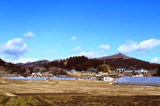 青空の下、左奥や右側にビニールハウスがあり、手前に広がる畑と、奥に木々に覆われた丘や山が見える風景の写真
