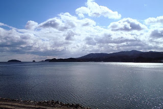 空には雲が広がり、穏やかな海と岩の多い海岸線、遠くに島と山々が見える、静かな海辺の風景の写真