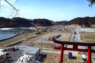 木々に覆われた小高い山を背景に、赤い鳥居が立つ道沿いに建物や工事資材が並ぶ開発中の風景の写真