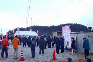 遠くには木々と建設機械が確認でき、周囲にはピンクと白色ののぼり旗やカラーコーンが配置されている献花式の会場で、集まった大勢の人たちが黙祷を行っている様子の写真