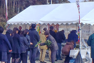 背景には木々が見える屋外で、のぼり旗が設置された白いテントの外で、配られた白い花を受け取っている列を作って並んだ人々の様子が写っている写真