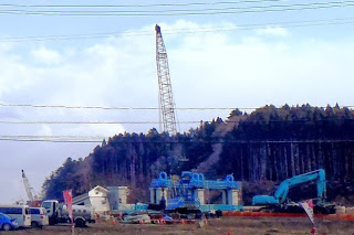 背景には木々が茂る丘が広がり、中央にアームを高く伸ばした大きなクレーンや青色のショベルカー等の建設車両が並ぶ工事現場の様子が写っている写真