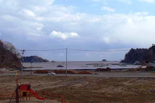 空は部分的に雲がかかる手前には赤いすべり台のある遊具が設置された遊び場があり、両側に陸地が広がる海辺の風景が写っており、水面には複数の船が浮かんでいる風景の写真