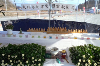 「東日本大震災慰霊献花式」と書かれたテントの下に、慰霊のための祭壇が設けられ、テーブルの上には小さな木製の像が並び、色とりどりの小さなキャンドルが置かれ、献花台には白い花が積まれている慰霊献花式の祭壇の写真