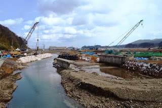 左岸と右岸にはそれぞれクレーンがあり、周囲には土砂や建設資材が積まれ、背景には山や建物が見える川の両岸で建設作業が行われている様子の写真