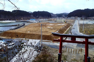 山々に囲まれた広い土地の風景で、中央には茶色い土が盛られた敷地が広がり周囲には道路や建物があり、左側には川や水辺、右手前には赤い鳥居が立っている写真