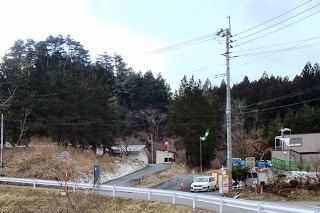 山間の自然豊かな環境の中にある舗装された道路の風景で、右側には小さな建物と白い車が駐車されており、左側には木々が茂り、電柱と電線が見え背景には高い木々が広がっている写真