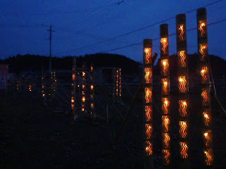 夜の空の下、等間隔に並べられた波模様などがデザインされた竹灯篭に橙色の明かりが灯っている写真