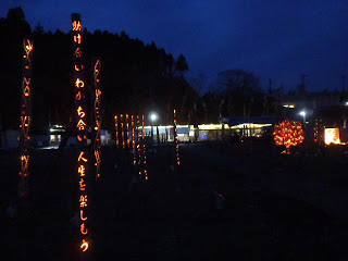 夜の広場で、明かりが灯った竹灯篭に彫られた、「助け合い わかち合い 人生を楽しもう」のメッセージがオレンジ色に浮かび上がっている写真