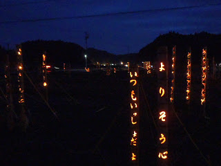 山影に囲まれた暗い広場に、複数の竹灯篭が立ち並び、「よりそう心」「いつもいっしょだよ」などのメッセージがオレンジ色に浮かび上がっている様子の写真