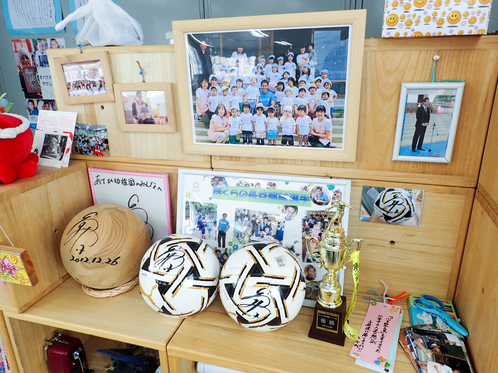木製の棚の上に、サイン入りのサッカーボールや木製ボール、優勝トロフィー、子どもたちと撮った集合写真、サッカー選手と思われる人物との記念写真、色紙や短冊が飾られており、幼稚園の教室内にある記念品コーナーの写真