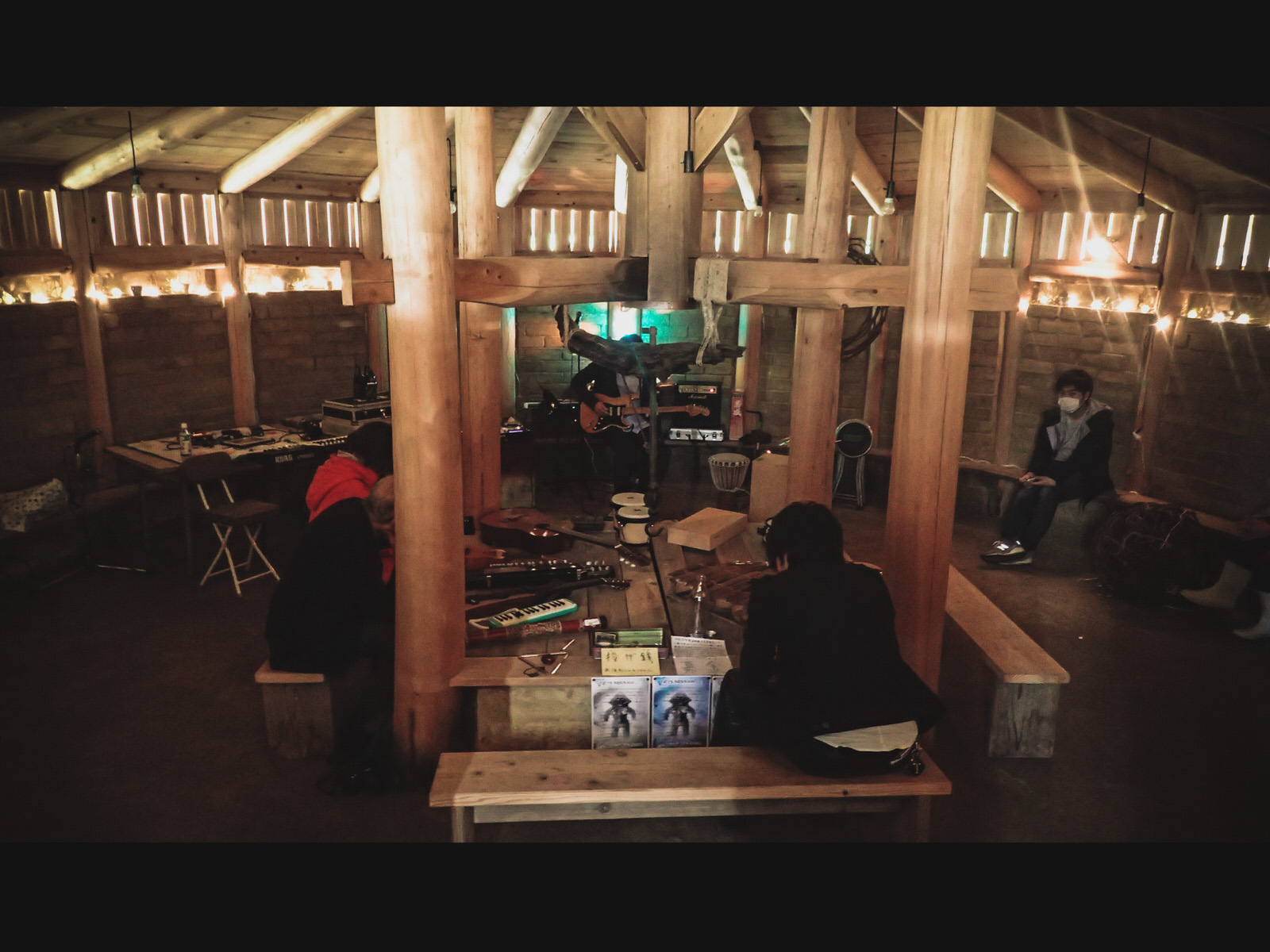 中央に4本の丸い木の柱がある、木造の円形の室内の中央や壁際にベンチが置かれ、演奏する人々やベンチに座る人々がいる様子の写真