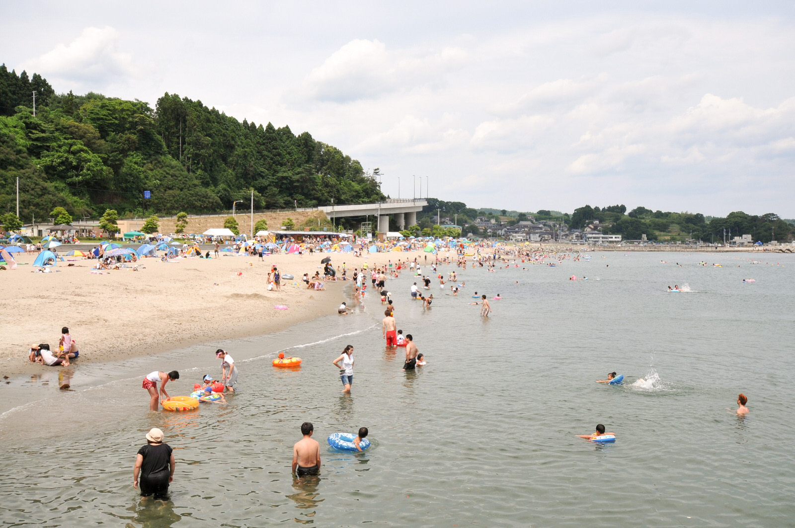 夏の晴れた日、砂浜と海に大勢の人々が集まり、カラフルなテントや浮き輪を使って海水浴やレジャーを楽しんでいる写真
