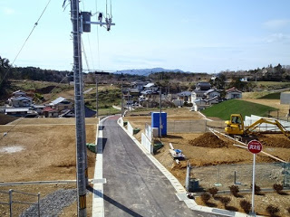 電柱や標識も設置され、舗装された道路を挟み両側で区画整備された宅地が整然と並び、右側では重機が作業を行っている工事中の写真