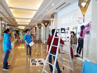 ホテルの広い屋内のロビーで、はしごや椅子を使って壁に写真や紙を貼り付けながら、数人の若者たちが展示準備をしている様子の写真