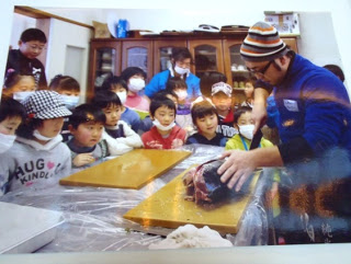 ニット帽をかぶった男性が教室内の調理台の上で大きな魚を包丁でさばいていて、その周りを多くの子どもたちが興味深そうに囲んで見つめている、お魚教室の様子の写真。