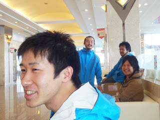 青いジャンパーを着た男性たちがソファに座ったり立ったりしながら談笑していて、手前には笑顔の男性が大きく写っている様子の写真