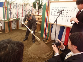 周囲で出席者が見守ったり、カメラで撮影しているなか、スーツ姿の男性が儀式の一環として砂の山に鍬を入れている様子の写真