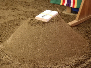 安全祈願祭で使われた円錐型の砂山の上に、奉納物として中央に文字が記載された長方形の白い封筒の様な物が置かれている写真