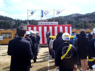 ステージが紅白幕で覆われステージの上には3つの旗が掲げられており並んで話を聞いている写真