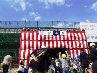 緑色の足場で覆われた建物の紅白幕の入り口下で子供たちが上を見て手をあげている写真