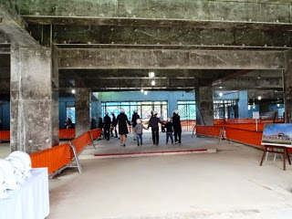 左側にはヘルメットが並べられたテーブルがあり、右側には建物の完成予想図が描かれた看板、建設中の建物の内部を奥に向かって進んでいる数人の人々を後方から写した写真