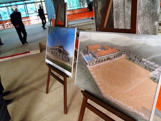 2つのイーゼルに置かれた建築図面で左側の図面は伝統的なデザインの建物を描いており、右側の図面は運動場や複数の建物を含む大きな複合施設が描かれている写真