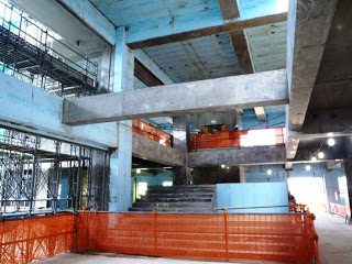 コンクリートの梁や柱が見え、階段が中央にありオレンジ色のフェンスが設置されており、安全対策が施されている建設中の建物の内部を写した写真