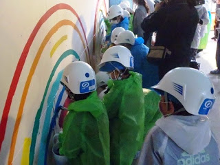 ヘルメットをかぶった子供たちが緑色や青色のレインコートを着て、壁に赤、オレンジ、黄色、緑、青の虹の絵を描いている様子を写した写真