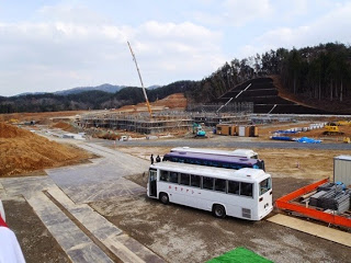 中央には足場が組まれた建物の骨組みがあり、その背後にはクレーンが見え手前の道路上には白いバスが停車している建設現場の写真