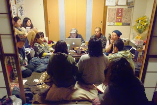 パソコンやお茶が置かれた和室のこたつを囲んで十人以上の人たちが座り、話し合いや作業をしている様子が写っている写真