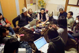 狭い和室の部屋に設置されたコタツの周りに多くの人たちが集まり、パソコンや資料を見ながら真剣に話し合いをしている様子の写真