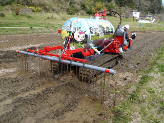 赤い車体に畑の写真やキャラクターがデザインされたタンクを乗せた液肥散布車が、後方に設置した長いパイプの穴から液肥を散布している様子の写真