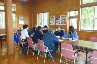 壁にポスターが貼られている木造の室内で、数人の参加者が机を囲んで話し合いをしている写真