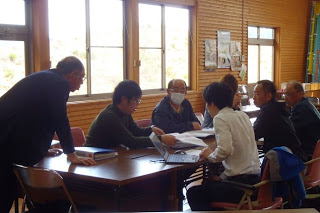 太陽光が差し込む窓が設置された木造の会議室で、7人の参加者が資料を手に話し合いをしている写真