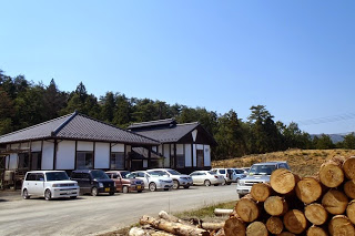 和風の平屋建物の前に多くの自動車が並び、右手前には伐採された丸太が積まれている森林組合の建物外観の写真